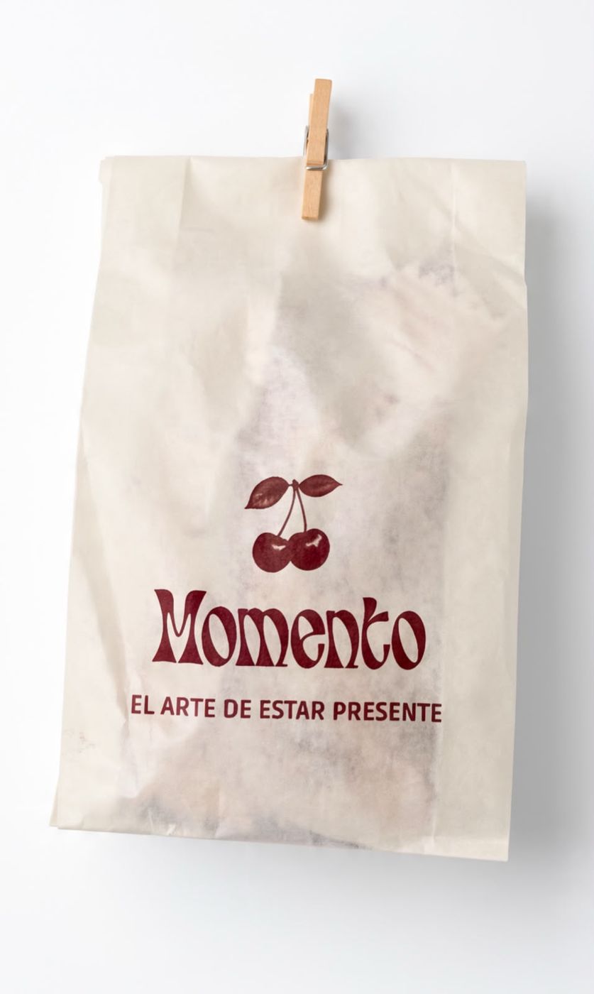 Bolsa de papel de Momento con la cereza, el logo y la frase «El arte de estar presente», cerrada con un perro de ropa.